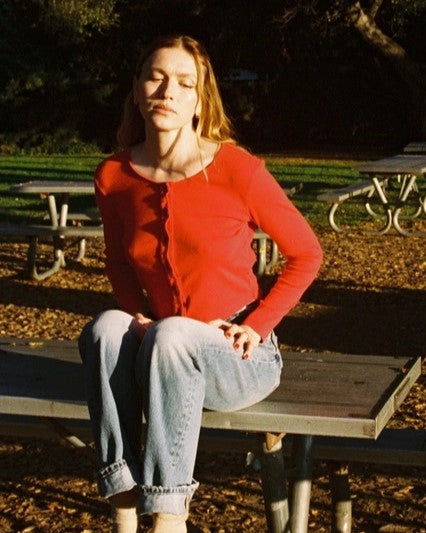 Bright red button-down long sleeve cardigan. Cropped and paired with high-wasited denim jeans rolled at the hem. Woman seated on picnic table in park.