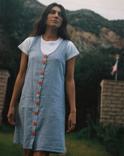 Button-down sleeveless washed linen sundress with navy stripes and bold red recycled buttons. Paired over a white short-sleeve tee with a mountain background.