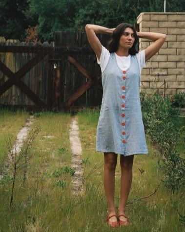 Button-down sleeveless washed linen sundress with navy stripes and bold red recycled buttons. Paired over a white short-sleeve tee with a mountain background.