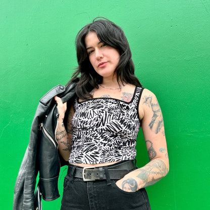 Black and white squiggle pattern zig zag tank top with thick straps, a square neckline, and ruche bodice detail. Paired with a black moto jacket and high-waisted denim. Made by woman-owned brand Nooworks in California. Bright green background.