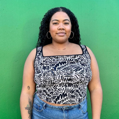 Black and white squiggle pattern zig zag tank top with thick straps, a square neckline, and ruche bodice detail. Paired with a black moto jacket and high-waisted denim. Made by woman-owned brand Nooworks in California. Bright green background.