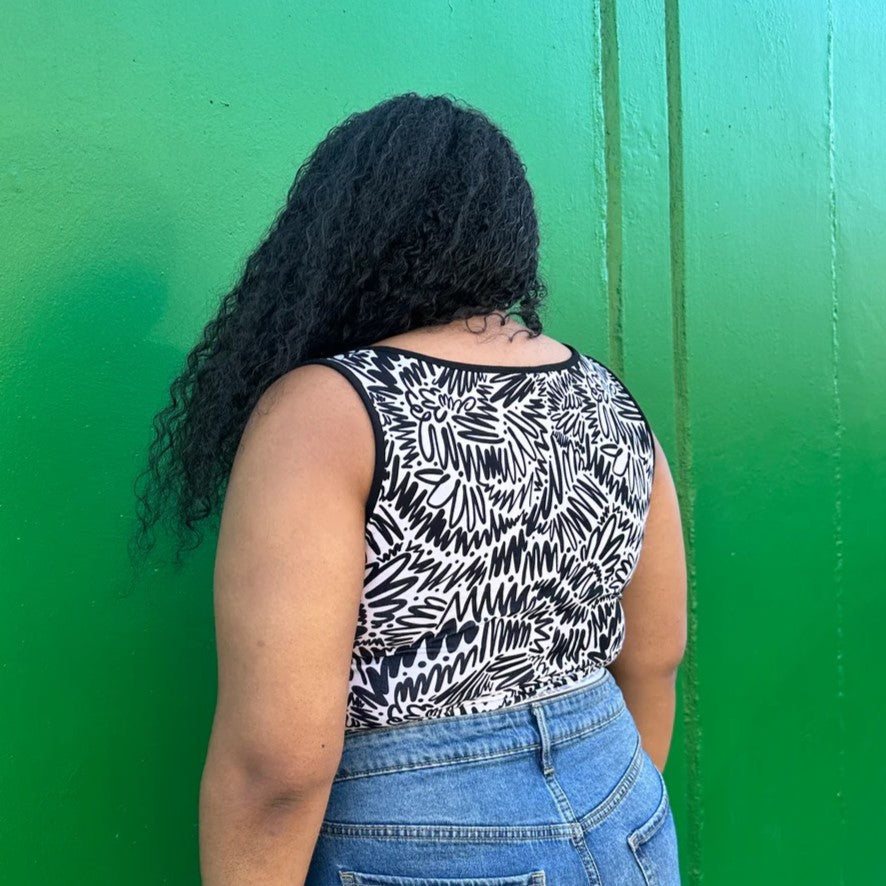 Black and white squiggle pattern zig zag tank top with thick straps, a square neckline, and ruche bodice detail. Paired with a black moto jacket and high-waisted denim. Made by woman-owned brand Nooworks in California. Bright green background.