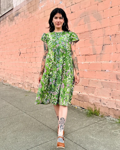 A green floral meadow dress with cap sleeves and a tiered skirt with flowers. Made in California by women-owned business Nooworks. Paired with clogs and large hoop earrings.