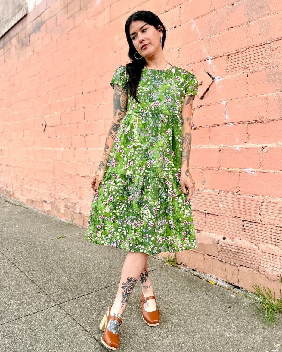 A green floral meadow dress with cap sleeves and a tiered skirt with flowers. Made in California by women-owned business Nooworks. Paired with clogs and hoops.