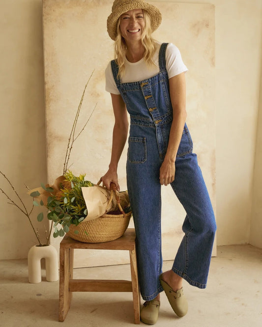 A thick-strapped sleeveless blue denim jumpsuit with pockets and a button-down front. Wide legs and a fitted waist.